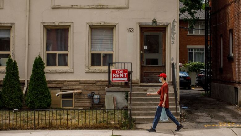 A home with a for rent sign on the front porch is pictured from the street at a pedestrian walks by.