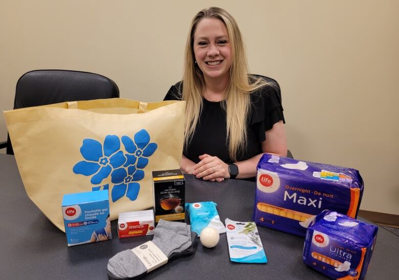 A woman with blond hair sits at a table, smiling. On the table are hot and cold compresses, socks, tea, Tylenol, panty liners, pads and a bath bomb.