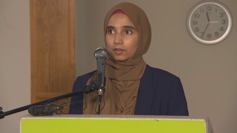 A woman wearing a brown hijab speaks at a microphone.