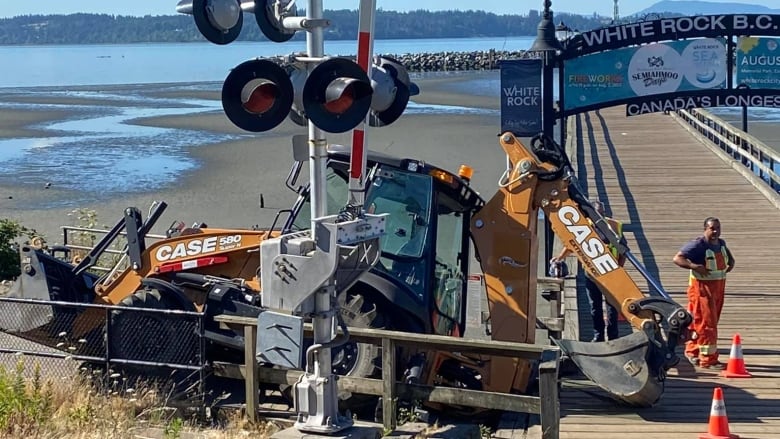 A yellow backhoe sits precariously on the entrance to a long pier.