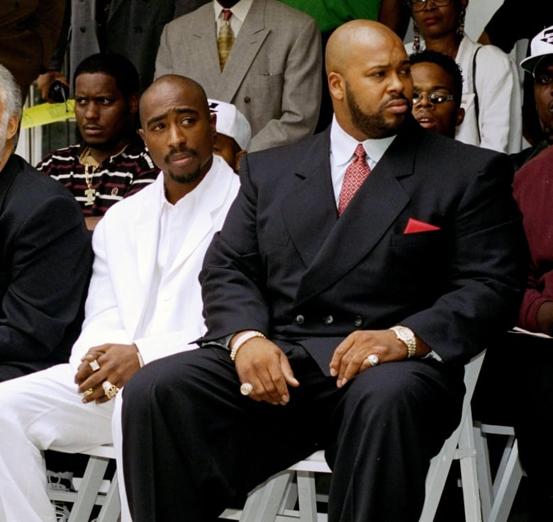 Two men are shown seated at an event, one wearing all-white and another in a suit and tie.