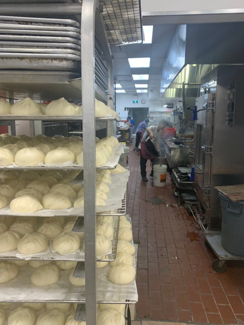 Trays of bao buns are shown on a metal rack in a commercial kitchen, with a worker in the background.