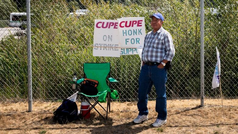 A man stands by a camping chair and picket signs for a bus driver's strike in front of a chain link fence.