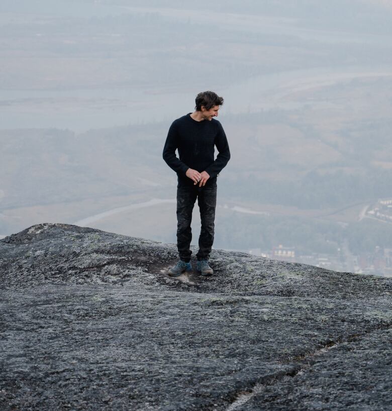 A slim man wearing all black stands at the top of a mountain.