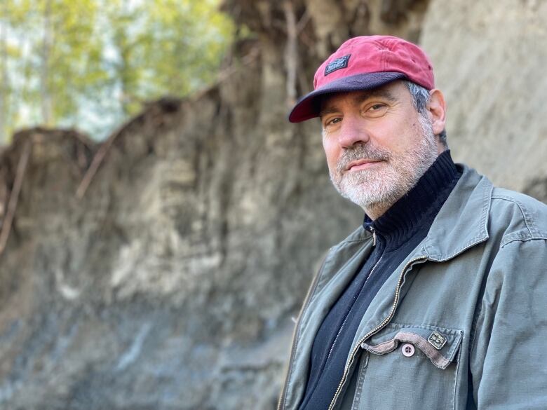 A bearded man in a cap stands in front of a steep embankment.