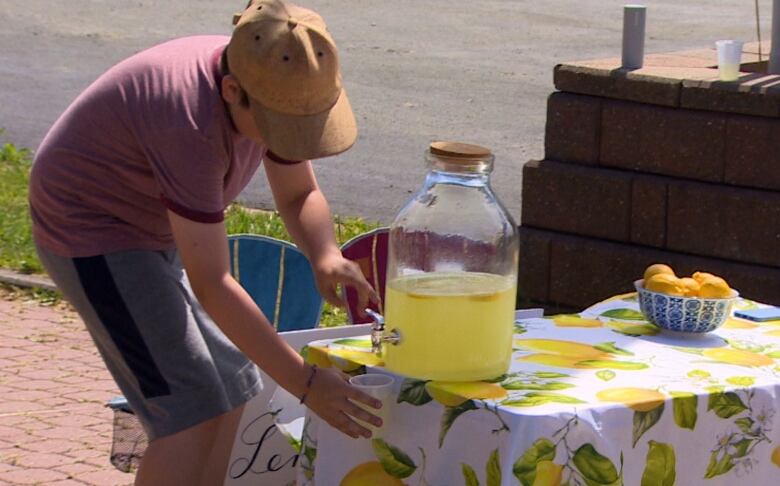 A boy wearing a red shirt and brown hat pours lemonade into a cup. 