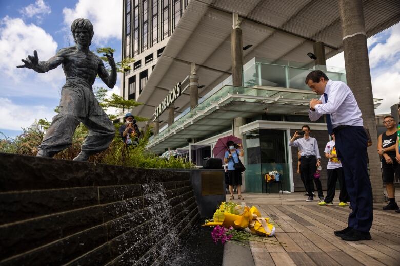 A man in a collared shirt and tie makes a praying motion before a statue, with flowers lain at the base of a monument.