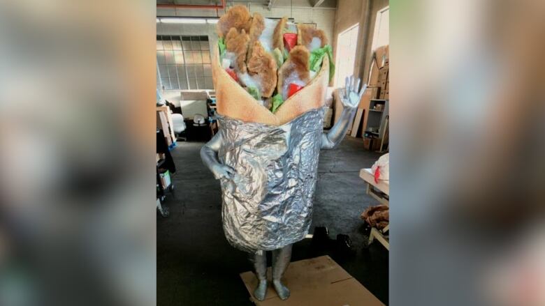 A person stands and waves dressed in donair costume, featuring tin foil, meat, white sauce, tomatoes and onion.