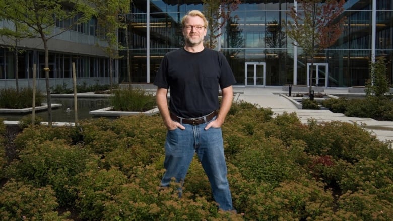 A man in a dark t-shirt and jeans stands in bushes in front of a glass building and courtyard.