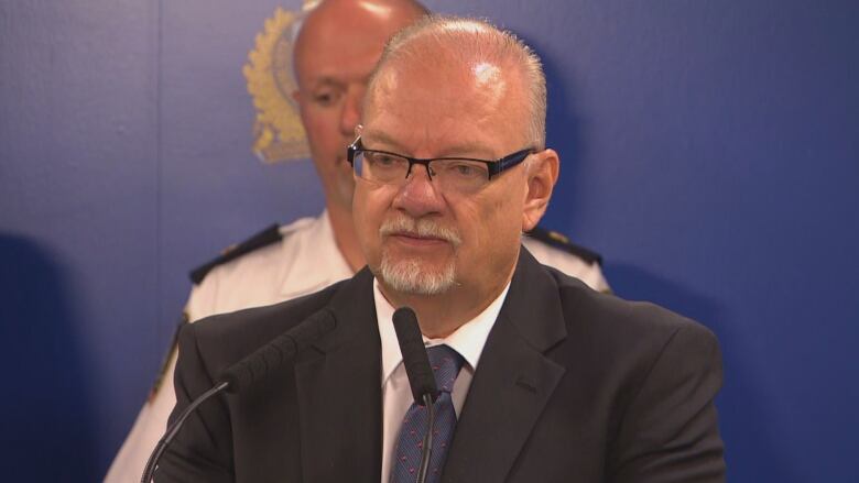 A man with a white goatee, glasses and short white hairs speaks at a podium