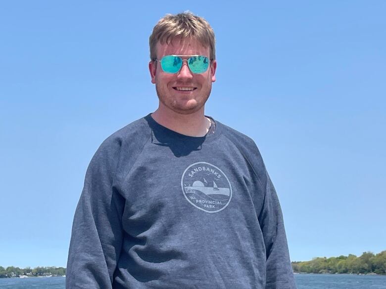 A man wearing a blue sweater and blue sunglasses poses for a photo by the lake on a sunny day