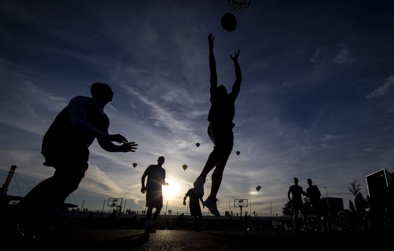 Silhouettes of boys playing basketball at sunset with clouds and silhouettes of hot air balloons in the background