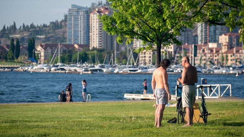 Two men naked on top stand on a grassland near a waterfront, with buildings in the background.