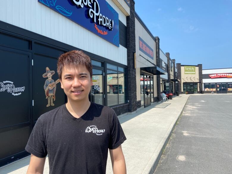 A man in a black shirt stands in front of a commercial plaza.