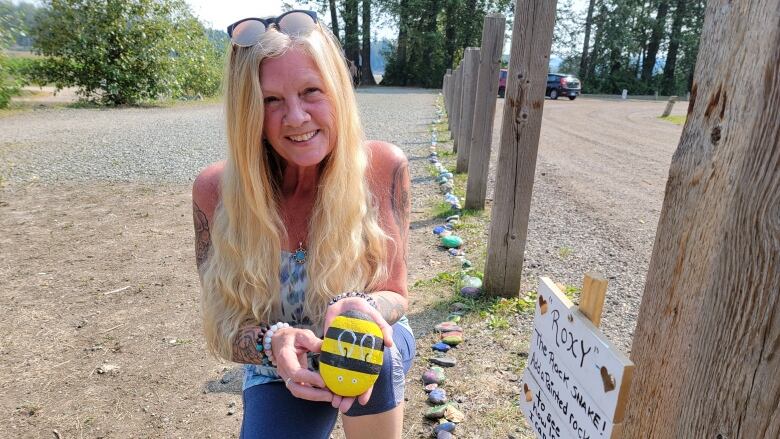 A woman holds up a rock painted to look like a bumble bee, with colourful rocks stretching behind her into the distance.