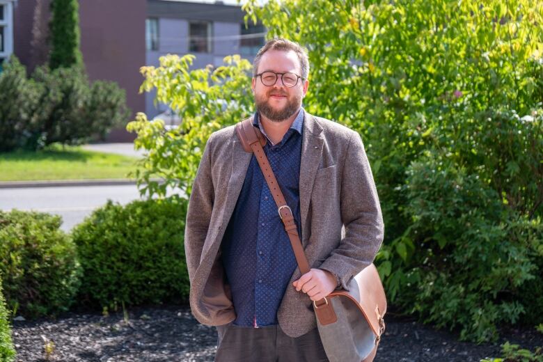 A man stands for a photo in front of a green bush, wearing a light brown jacket, a blue shirt, and a bag