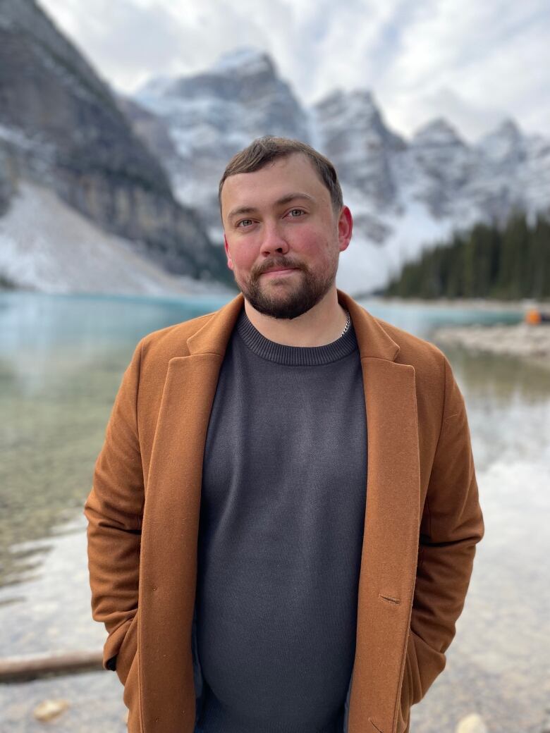 A man in a camel brown coat stands in front of a valley.