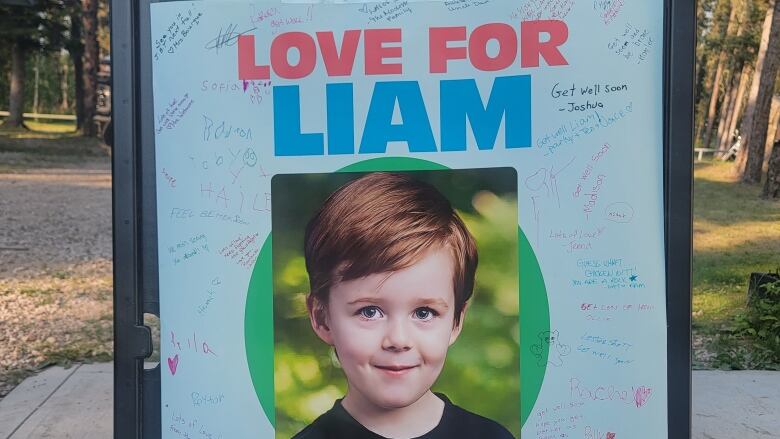 A sign stands in a park with a photo of a young boy, and the words, 'Love For Liam.'