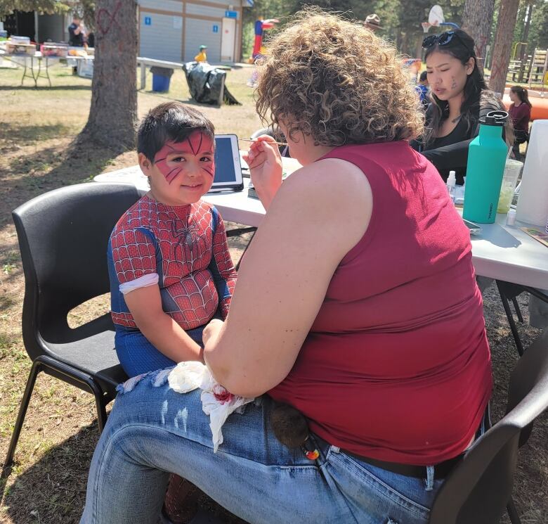 A small boy in a Spiderman suit gets his face painted at a park.