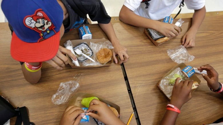 The hands of four young students are seen from above as they eat boxed lunches at a school dining table.