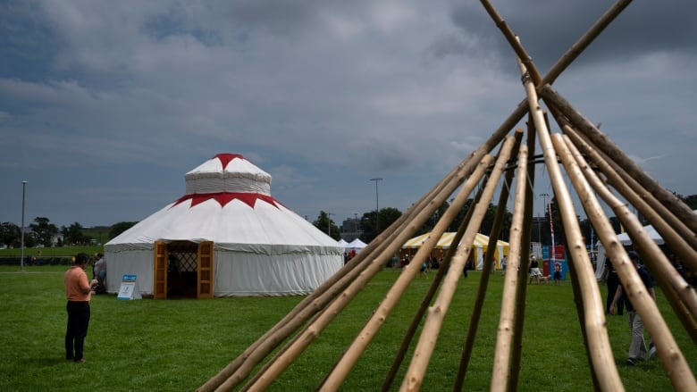 A man stands outside a ceremonial tent. A wooden teepee framework is in the foreground.