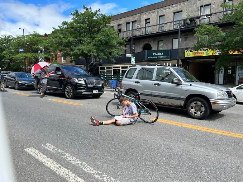 A boy is seated on the road beside his parked bike as cars drive on the adjoining lane. The boy is reading a book. He is participating in the human bike chain formed along Montreal's Parc Avenue on July 22, 2023.