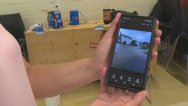 A woman holds a cellphone showing a picture of her home, the driveway is completely flooded.