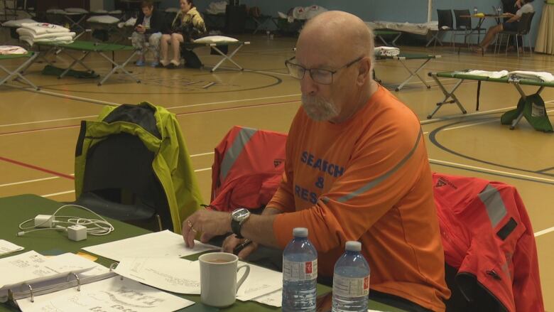 A man in an orange Search & Rescue t-shirt sits at a table going over paperwork. Behind him are several cots, some with people sitting on them.