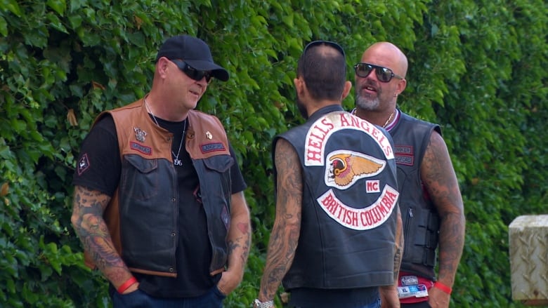 Three men with tattoos and leather vests on speak with one another.