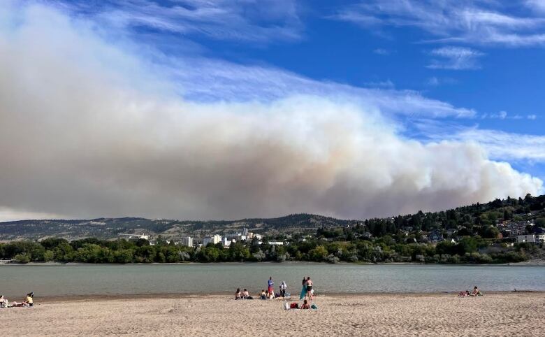 People in the distance along a long lakeshore watch a vast, long plume of white smoke billow over the lake from behind forested hills.