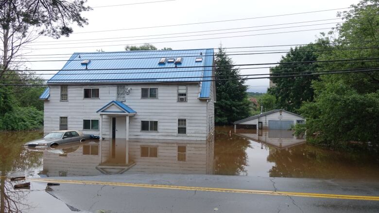 A large white building is seen surrounded by a water, flooded. A small sedan sits in front of the house and is also flooded.