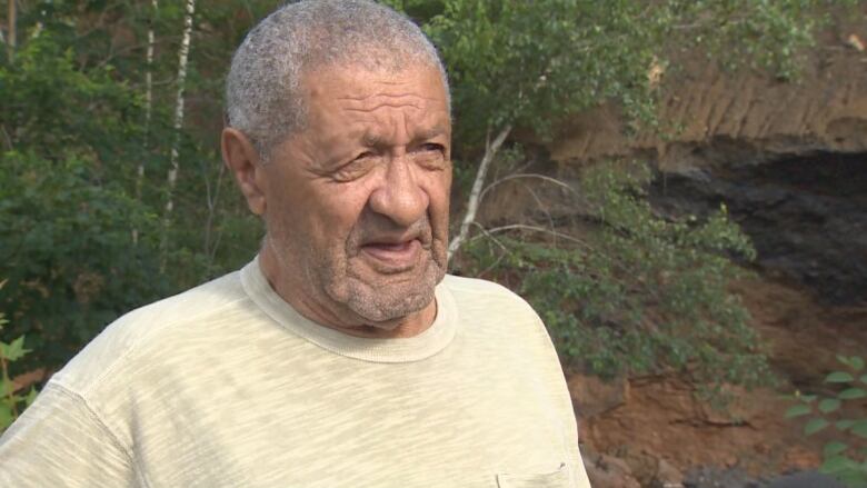 A man in an ecru t-shirt speaks to the camera.