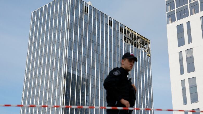 A man in a black uniform and cap stands behind a police tape. In the background is a highrise building showing broken windows and damage to the top floors.