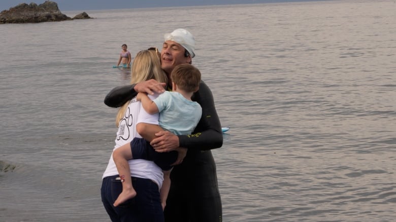 A man in a wetsuit is pictured hugging a woman and young child with the ocean in the background. 