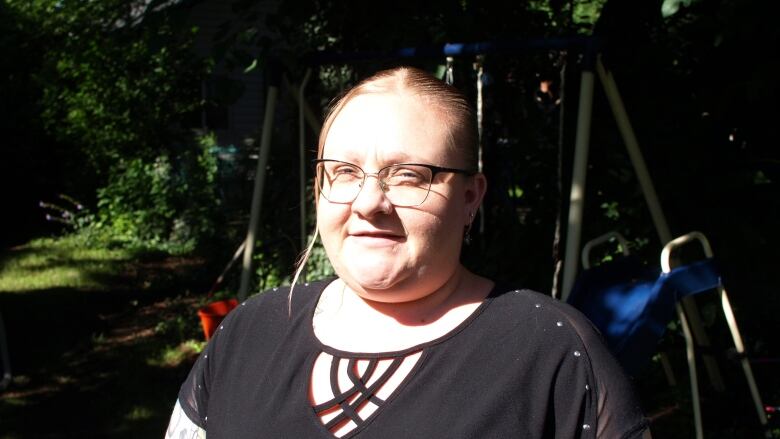 A person wearing glasses stands in a wooded area in front of a children's playset.