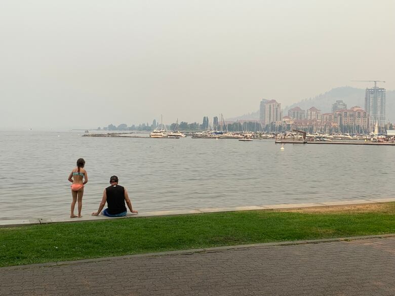 Two people sit on the shore of a large lake directly across from a city skyline partially obscured by haze.