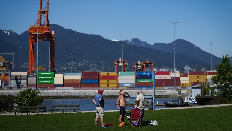 Three people stand by the Vancouver docks with containers behind them, with the ocean and mountains as a background. One of the drinks from a can.