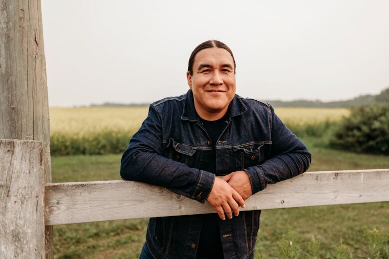 A man in a blue jean jacket leans on a wooden fence.