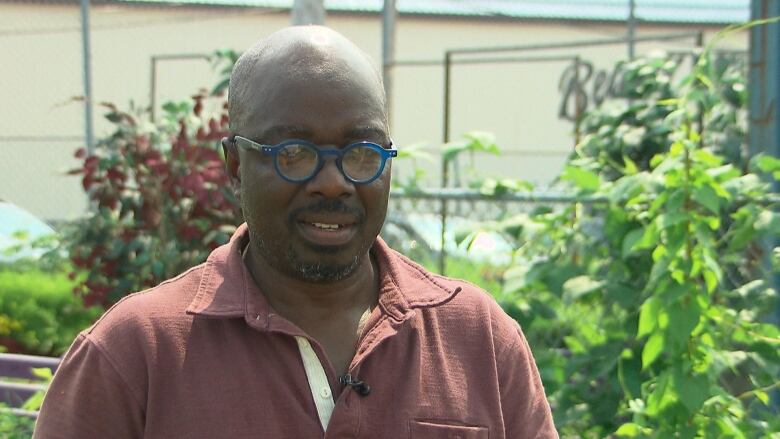 Bald man with blue-rimmed glasses stands in front of lush plants.