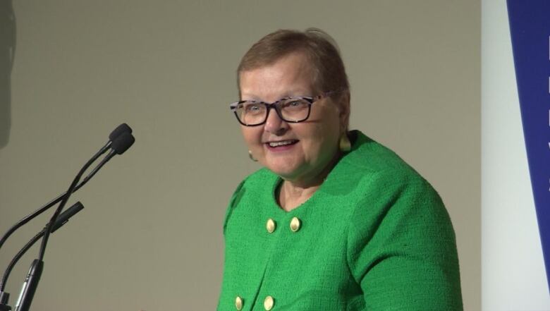 A spectacled woman in a green vest suit with golden buttons smiles.