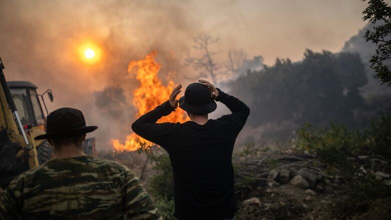 Two men pictured from behind watching a massive fire burning. One man has his hands on his head. 