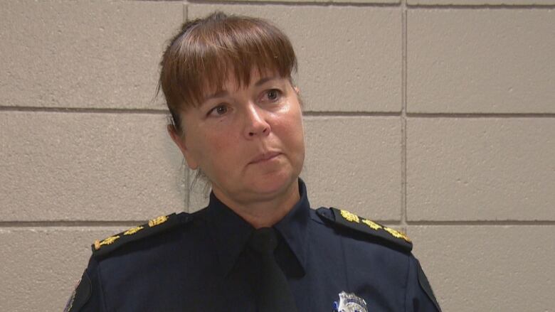 A white woman, with brown hair tied in a pony tail and bangs that cover her forehead, is wearing a navy blue police officer's uniform. She is standing in front of a brown, cement-block wall.