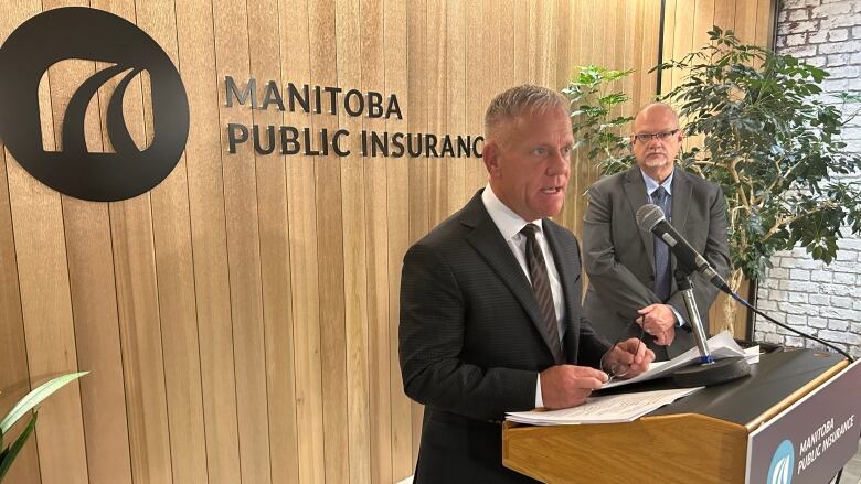 A man is shown speaking at a podium. Another man is seen standing at the side. Behind them is a sign that says 