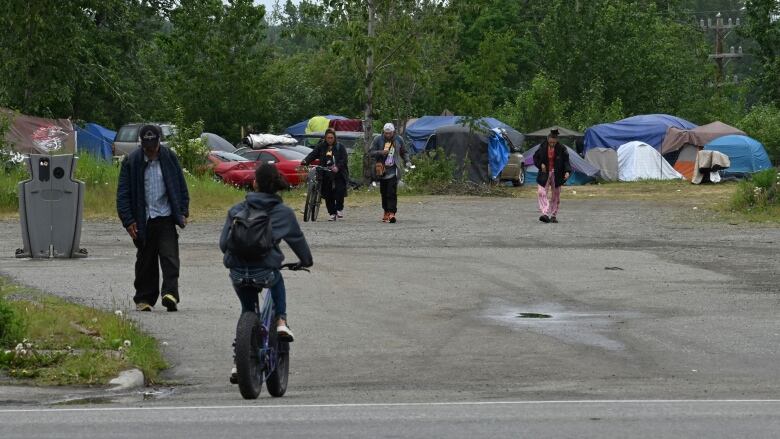 Some people walking or on bikes are seen coming and going from an area where some tents are set up. 