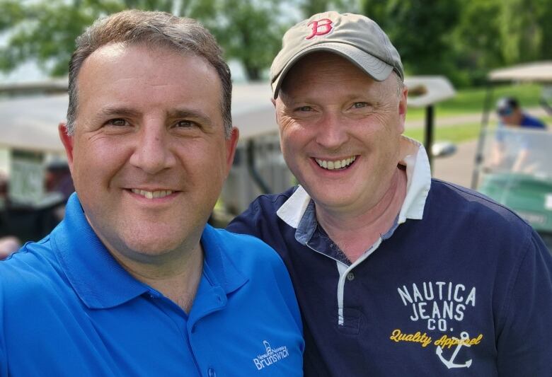 A selfie of two men side-by-side. Both are wearing blue and the man on the right has a baseball cap on.