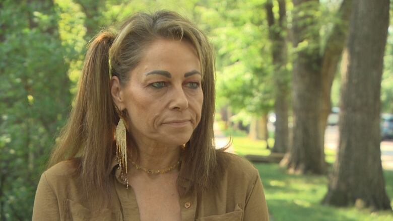 A woman with beaded earrings and pigtals looks serious as she talks to someone off-camera in a treed area.