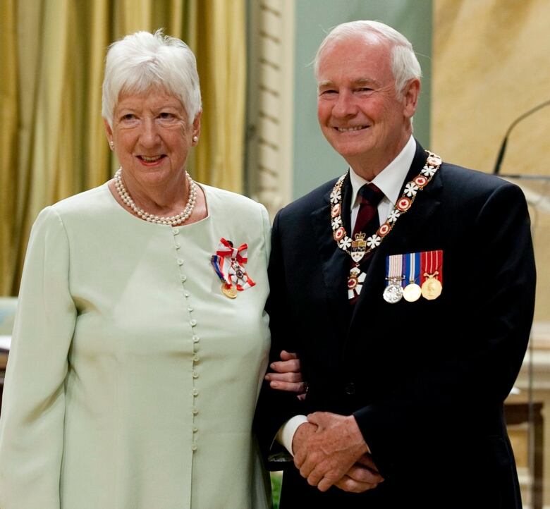 A woman with short white hair poses next to a balding man with white hair, who is wearing various medals.