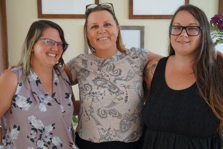 Three women stand next to each other. Two are wearing floral tops with the other is wearing a black shirt.