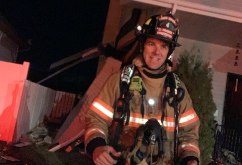 A man in full firefighting gear walks toward the camera in the dark. Reflective strips on his suit standout and the fence behind him glows red from the light. 