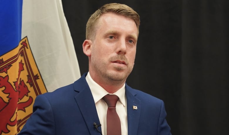 A man in a suit and tie stands in front of a flag.